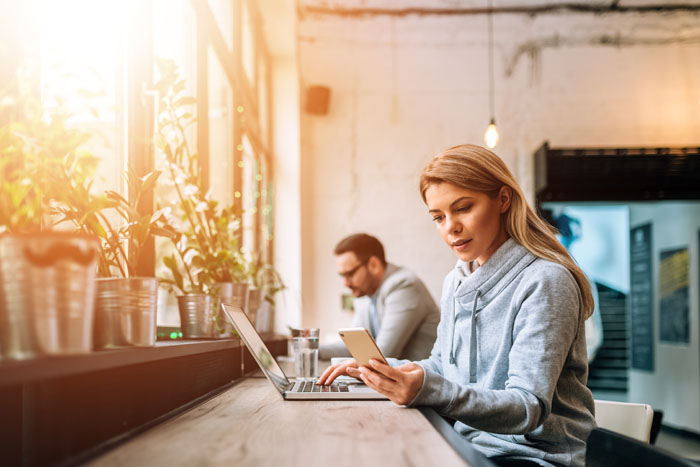 woman checks her smartphone as she works on a laptop in an office beside a coworker
